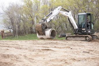 Bobcat E55 compact excavator (mini excavator) lifts drain pipe with clamp attachment. 